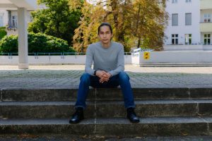 PHOTO 1 - Jay Afrisando, a medium-skin-toned male with bunned hair, sits on outdoor neighborhood concrete steps in a serious pose, with his fingers clasped and arms on his thighs. He wears dark blue jeans, a light gray sweatshirt, and black shoes. Photo: © DAAD Artists-in-Berlin Program / Diana Pfammatter.